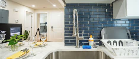 A modern kitchen with a stainless steel sink, a blue-tiled backsplash, and a dining area set with plates, glasses, and a plant.