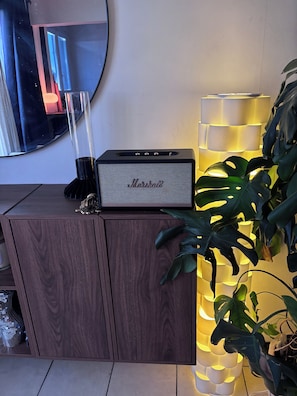 Un meuble en bois sur lequel est posé un amplificateur Marshall, un miroir et une plante, avec un luminaire vertical placé à côté.