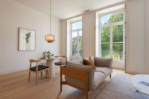 A modern living room with a sofa, a dining table, chairs, and a large window with a view of greenery. - Tripas-Coração: Mouzinho32 1st Floor Apt D (Porto)
