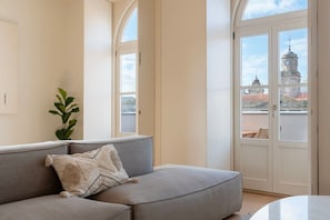 Ein Wohnzimmer mit grauem Sofa, einer Pflanze und Blick auf ein Gebäude mit einem Glockenturm durch eine Glastür.