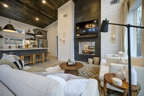 A modern living room with a fireplace, a kitchen with a bar area, and a large window. - Romantic Forest Escape | Hot Tub + Soaking Tub (Broken Bow)