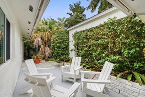 A covered patio with white chairs and a small table, surrounded by lush greenery and a white wall. - Upscale and Inviting Naples Home Near the Beach (Naples)