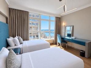 Ein Hotelzimmer mit zwei Betten, einem Schreibtisch, einem Stuhl, einem großen Fenster mit Blick auf Gebäude und Wasser sowie einer Deckenleuchte.