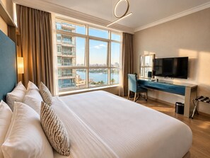 A hotel room with a large bed, a desk, a chair, a TV, and a view of a cityscape through large windows. - VESTA  - Executive Suite - 2BR - Partial Nile View (Cairo)