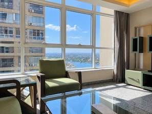 A modern hotel room with a large window, a green armchair, a glass coffee table, and a green cabinet. - VESTA  - Executive Suite - 2BR - Partial Nile View (Cairo)