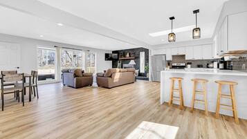Un salón amplio con piso de madera, un sofá, una mesa de comedor y una cocina con encimera blanca y taburetes de madera.
