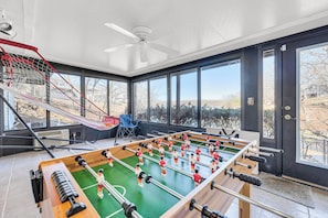 Una habitación con mesa de fútbol de mesa, ventilador de techo, ventanas amplias y vistas al exterior.