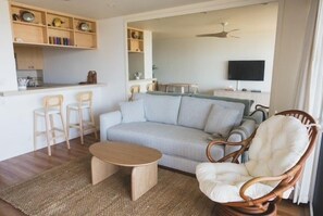 Ein Wohnzimmer mit einer Couch, einem Holzstuhl, einem kleinen Tisch und einer Küchenzeile im Hintergrund.