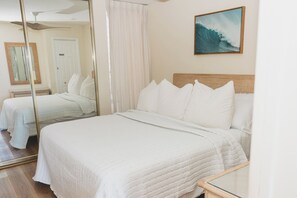A hotel room with a large bed, a mirror, a painting of a surfer, and a ceiling fan. - New unit to rent.  Adorable 1 bed/1 bath condo. (Lahaina)