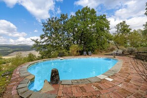 A swimming pool with a brick border, surrounded by trees and a view of the landscape.