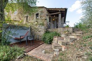 A rustic stone building with a wooden deck, outdoor seating, and a staircase leading to a covered area.
