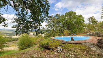 A swimming pool with a stone border and lounge chairs, surrounded by trees and a view of the countryside.