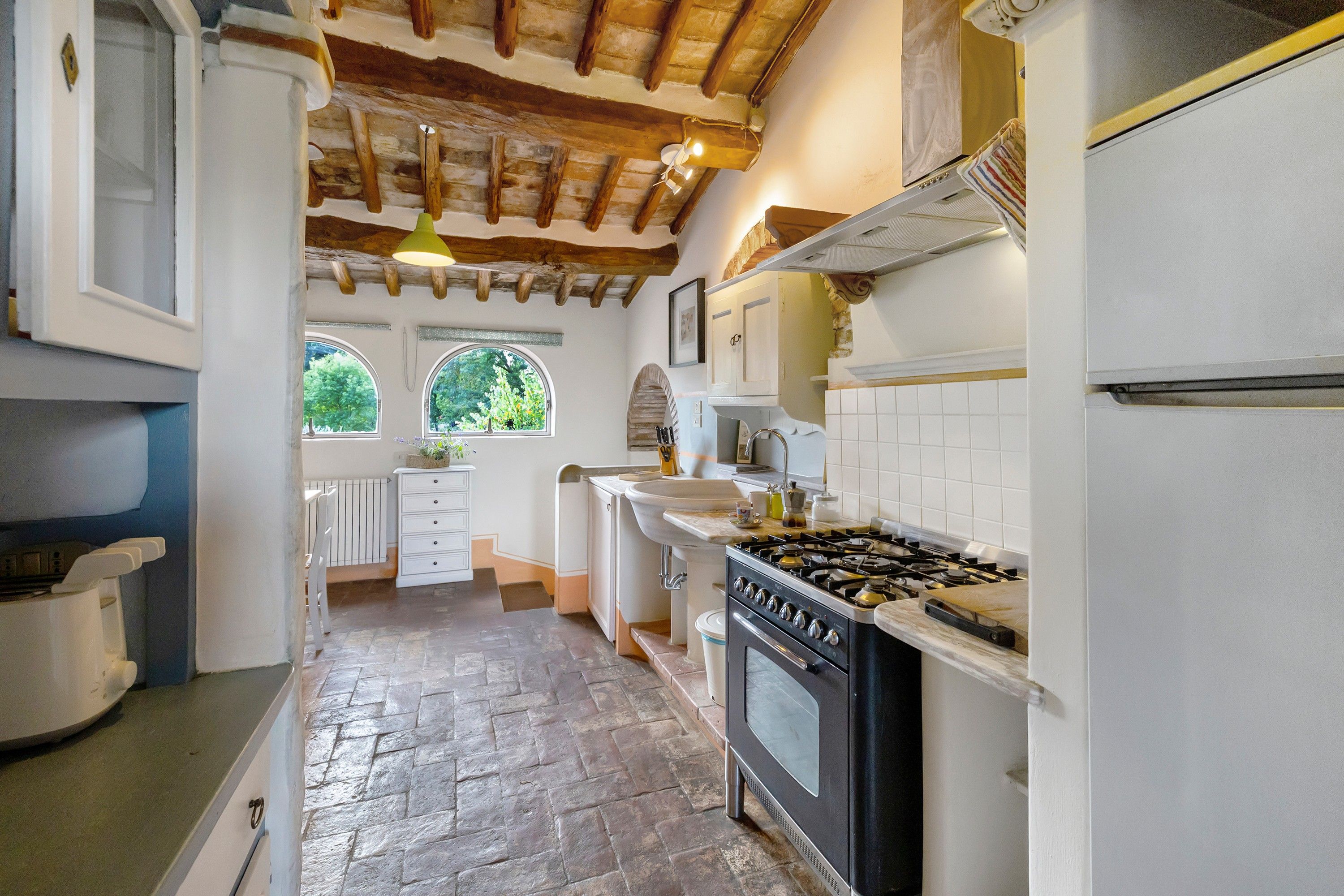 A kitchen with a stone floor, wooden beams, white cabinets, a stove, and a window with a view of trees.
