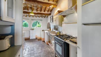 A kitchen with a stone floor, wooden beams, white cabinets, a stove, and a window with a view of trees.
