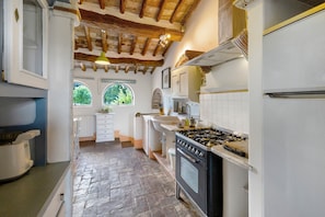 A kitchen with a stone floor, wooden beams, white cabinets, a stove, and a window with a view of trees.