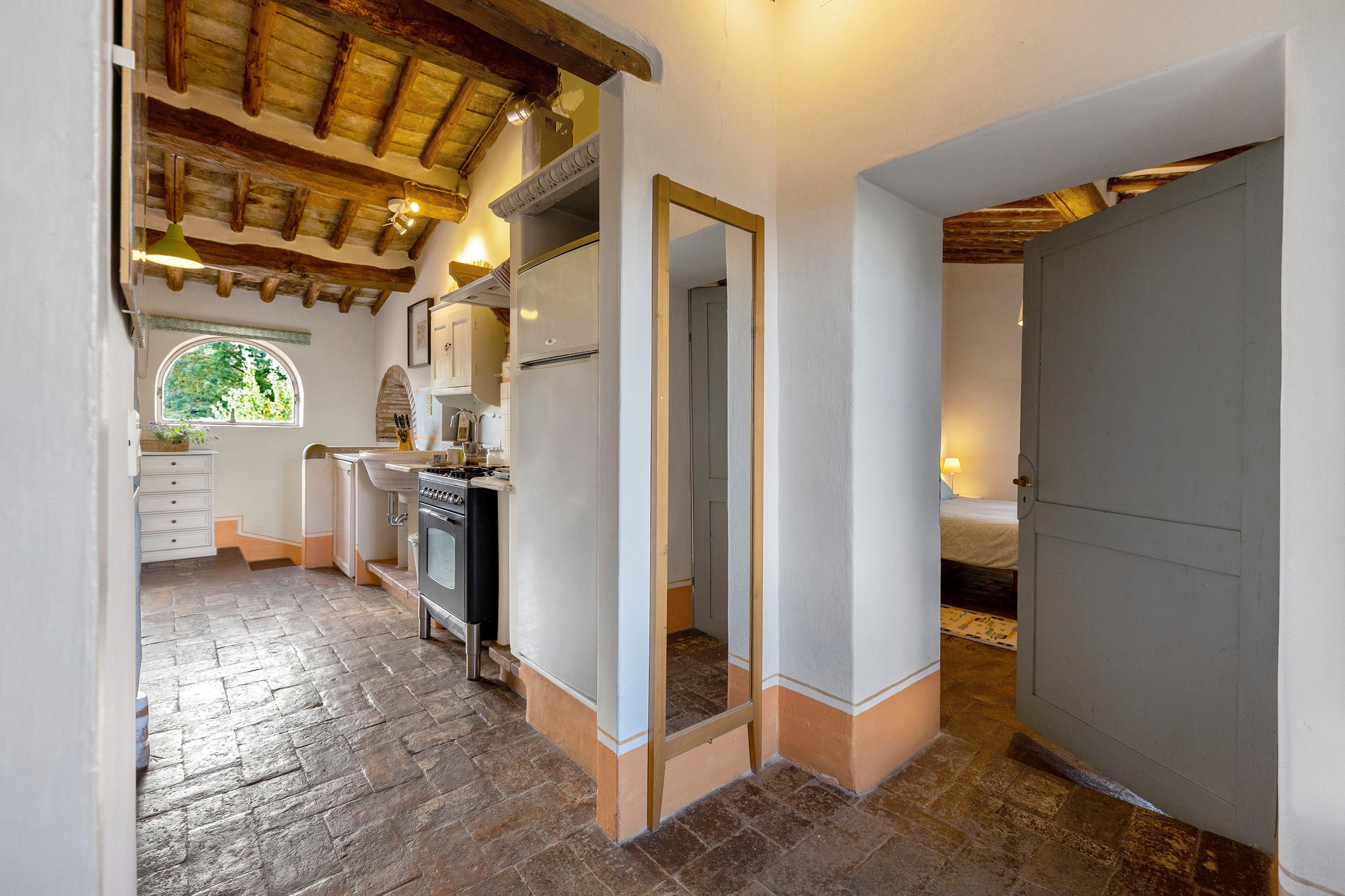 Een smalle gang met stenen vloer, houten plafond, een keuken met fornuis en spoelbak, en een slaapkamer met bed en nachtkastje.