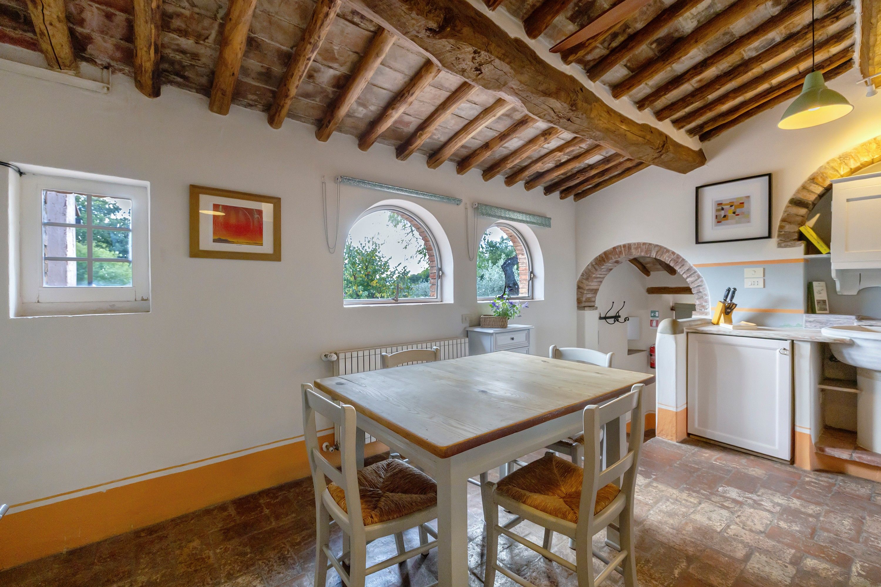 A rustic kitchen with a wooden table and chairs, a hanging light fixture, and a view through an arched window.