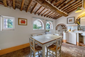 A rustic kitchen with a wooden table and chairs, a hanging light fixture, and a view through an arched window.