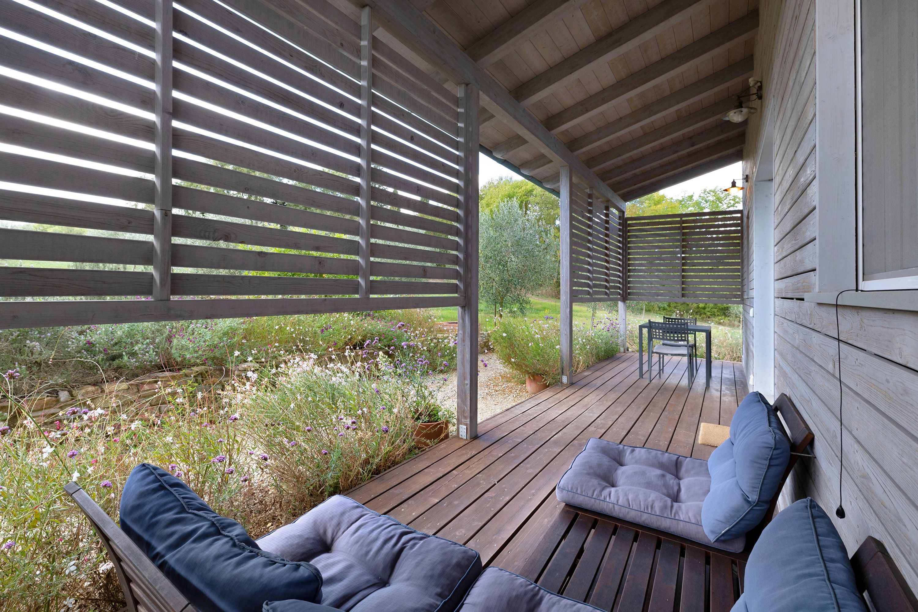 A covered outdoor seating area with wooden benches and cushions, a table, and a view of a garden with various plants and flowers.