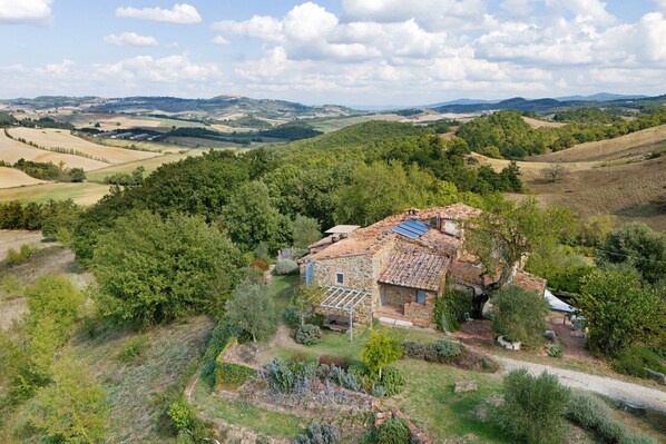 A stone house with a sloped roof, surrounded by lush greenery and fields.