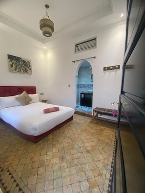 A hotel room with a bed, a small table, a bench, a bathroom with a shower, and a view through an archway. - Riad Alfombra (Marrakech)