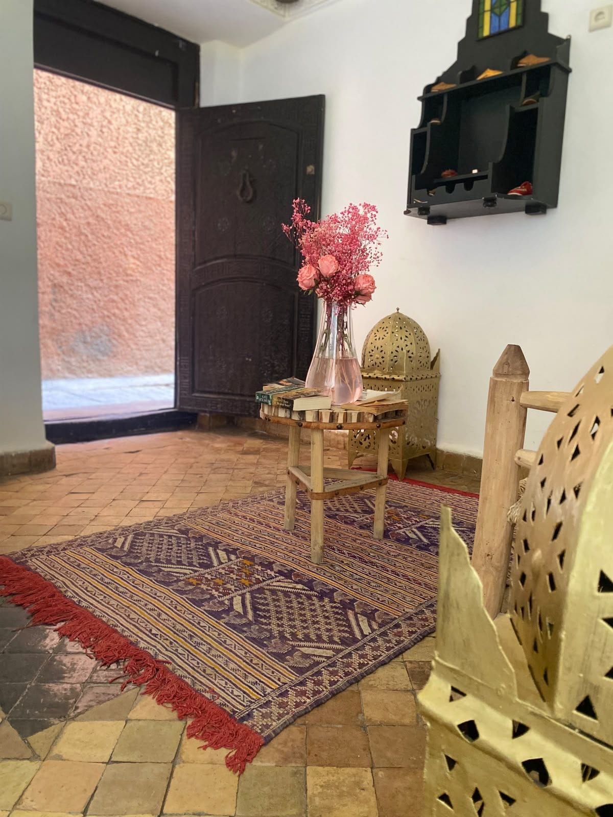 A room with a patterned rug, a wooden chair, a small table with a vase of flowers, and a decorative wall shelf.