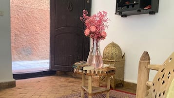 A room with a patterned rug, a wooden chair, a small table with a vase of flowers, and a decorative wall shelf.