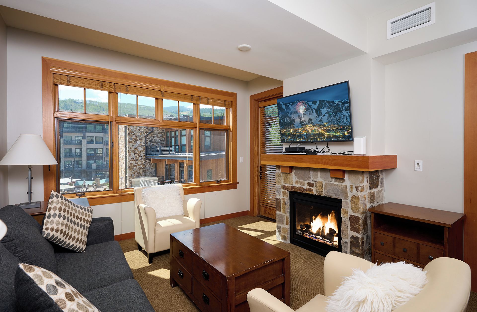 A living room with a stone fireplace, a television mounted above it, and a large window offering a city view.