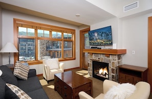 A living room with a stone fireplace, a television mounted above it, and a large window offering a city view. - Luxury 2 Bed, 2 Bath, Capitol Peak, In the heart of Base Village, steps to lift! (Snowmass Village)