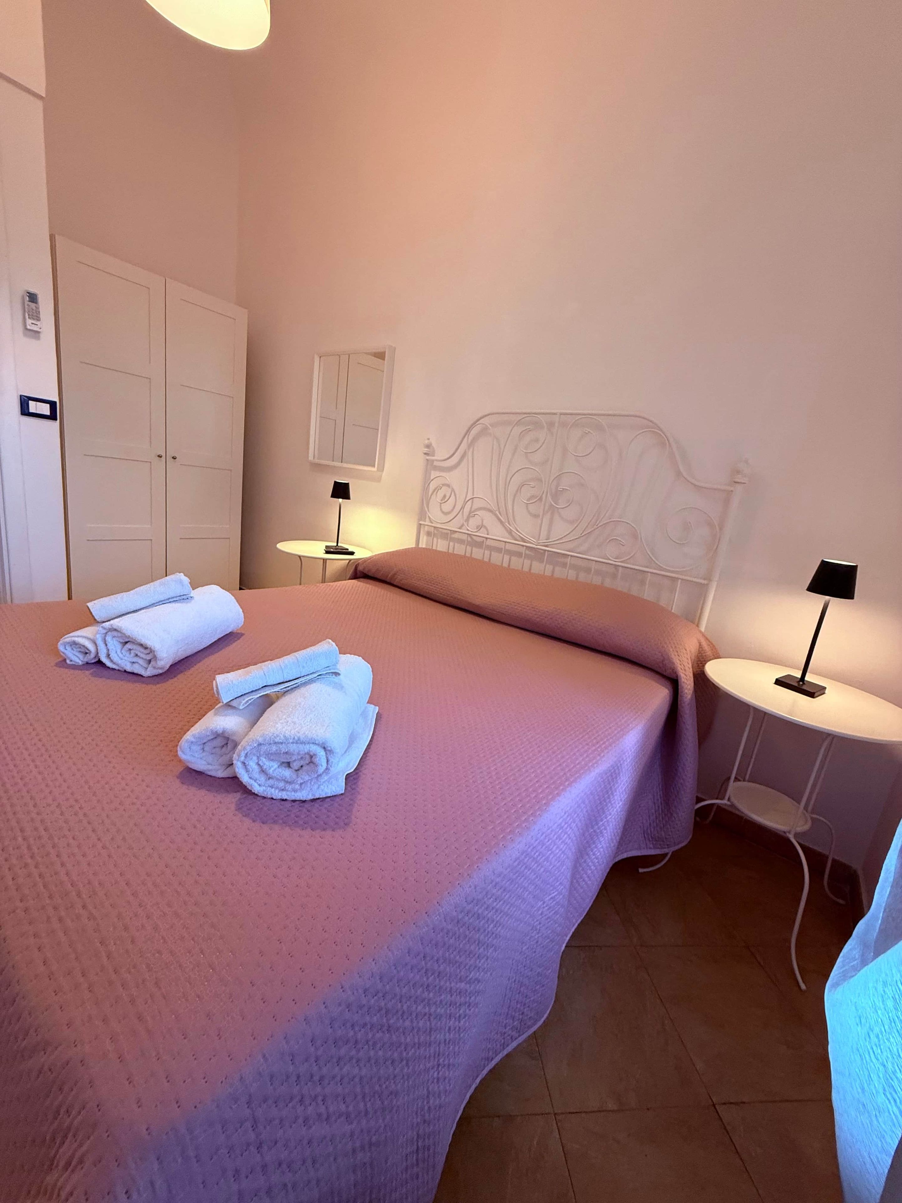Un lit bien fait, avec des serviettes pliées, une tête de lit en métal blanche et deux tables de chevet avec des lampes.