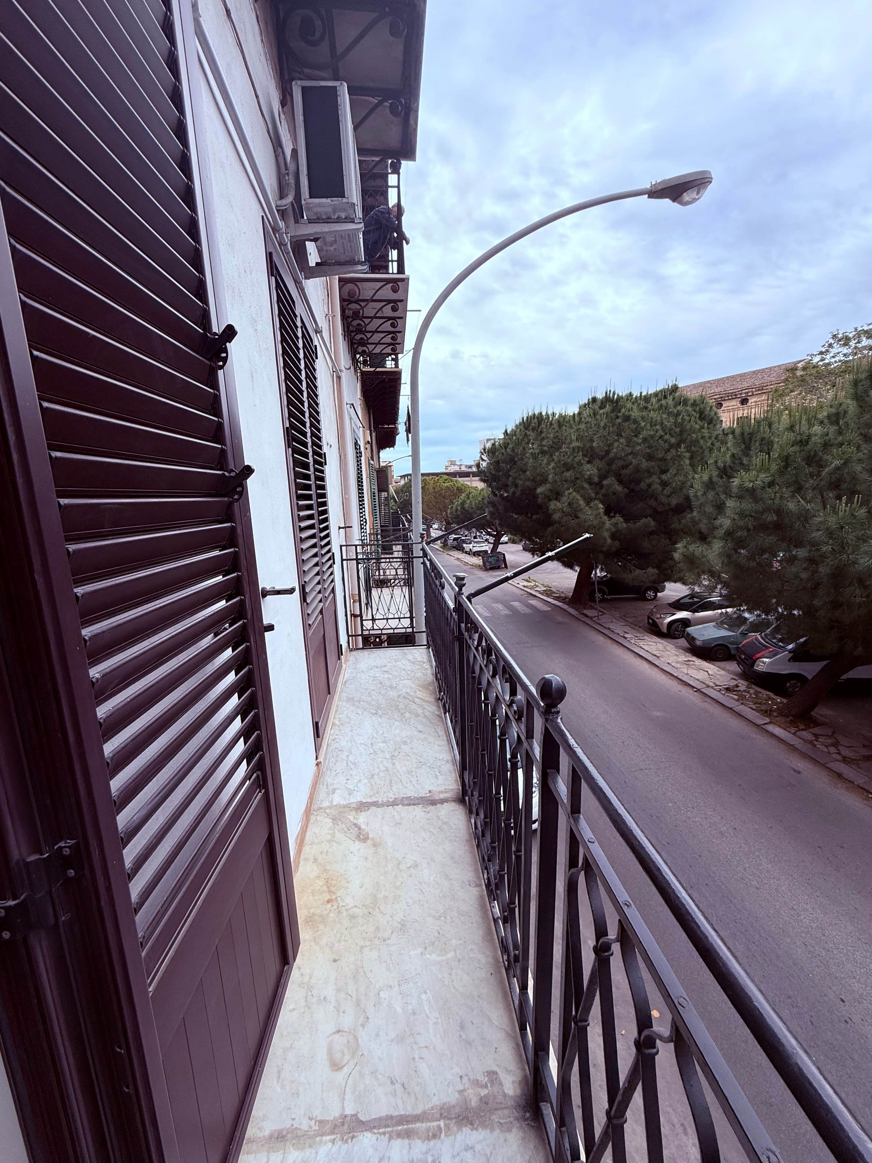 Ein schmaler Außenkorridor mit Metallgeländer, eine Straße mit parkenden Autos und ein klarer Himmel.