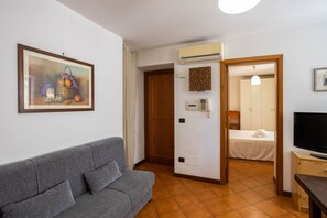 Un salon avec un canapé gris, un tableau représentant des fruits, un appareil de climatisation, une porte donnant sur une chambre à coucher et un téléviseur posé sur un meuble en bois.