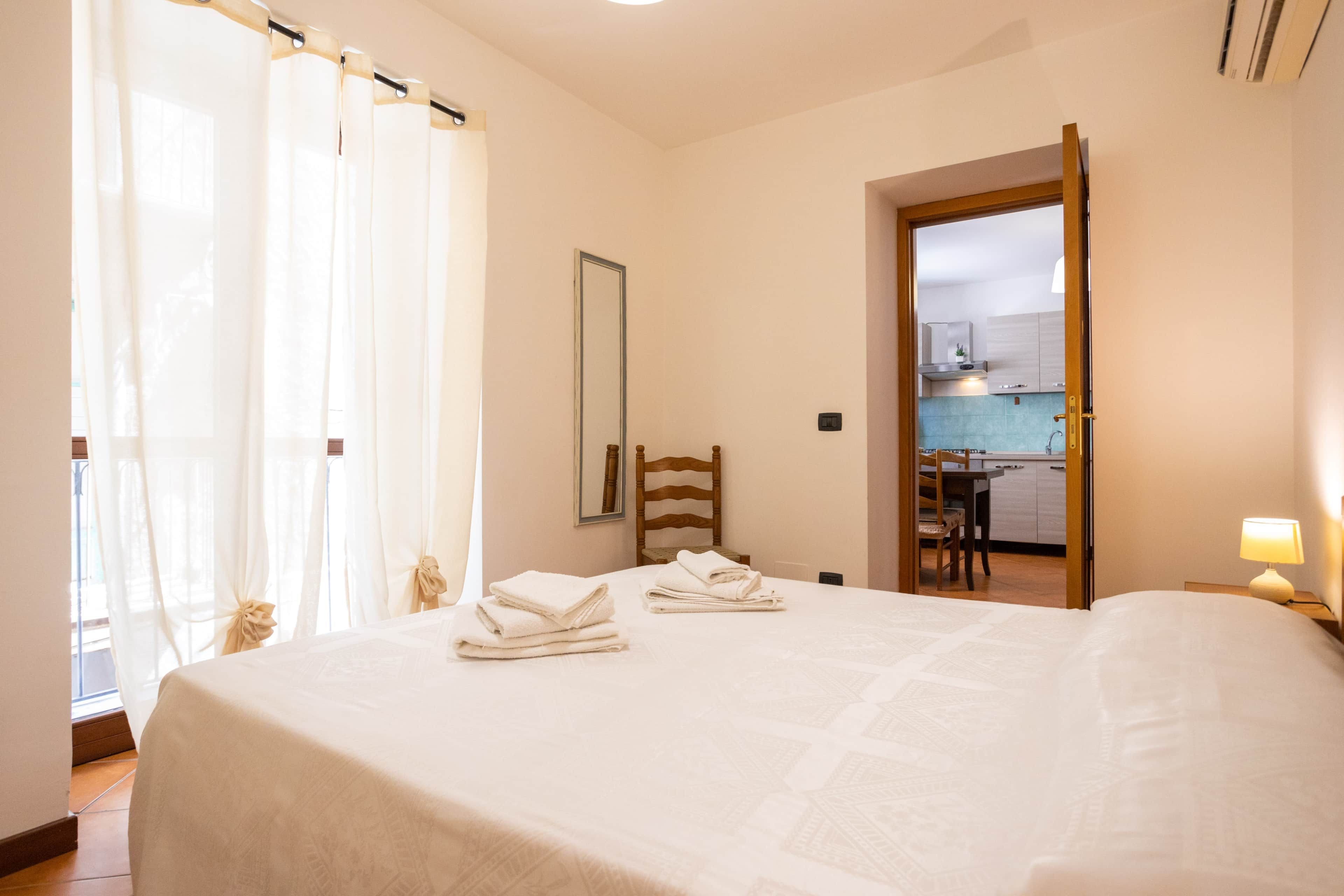 Une chambre d’hôtel avec un lit, une chaise en bois, un miroir et une vue sur une cuisine par une porte ouverte.
