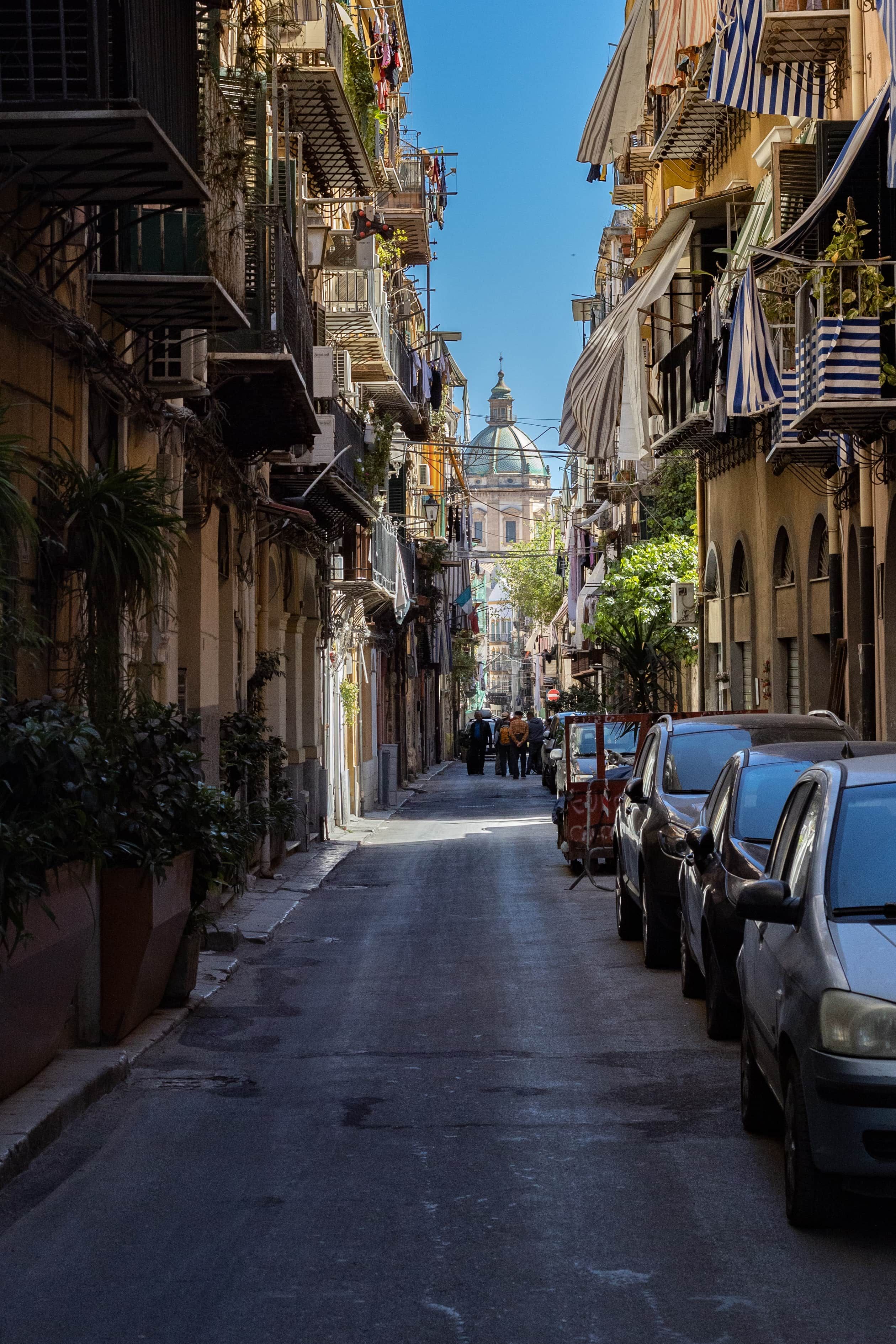 Une rue étroite bordée d’immeubles à plusieurs étages, de voitures garées et de quelques piétons.
