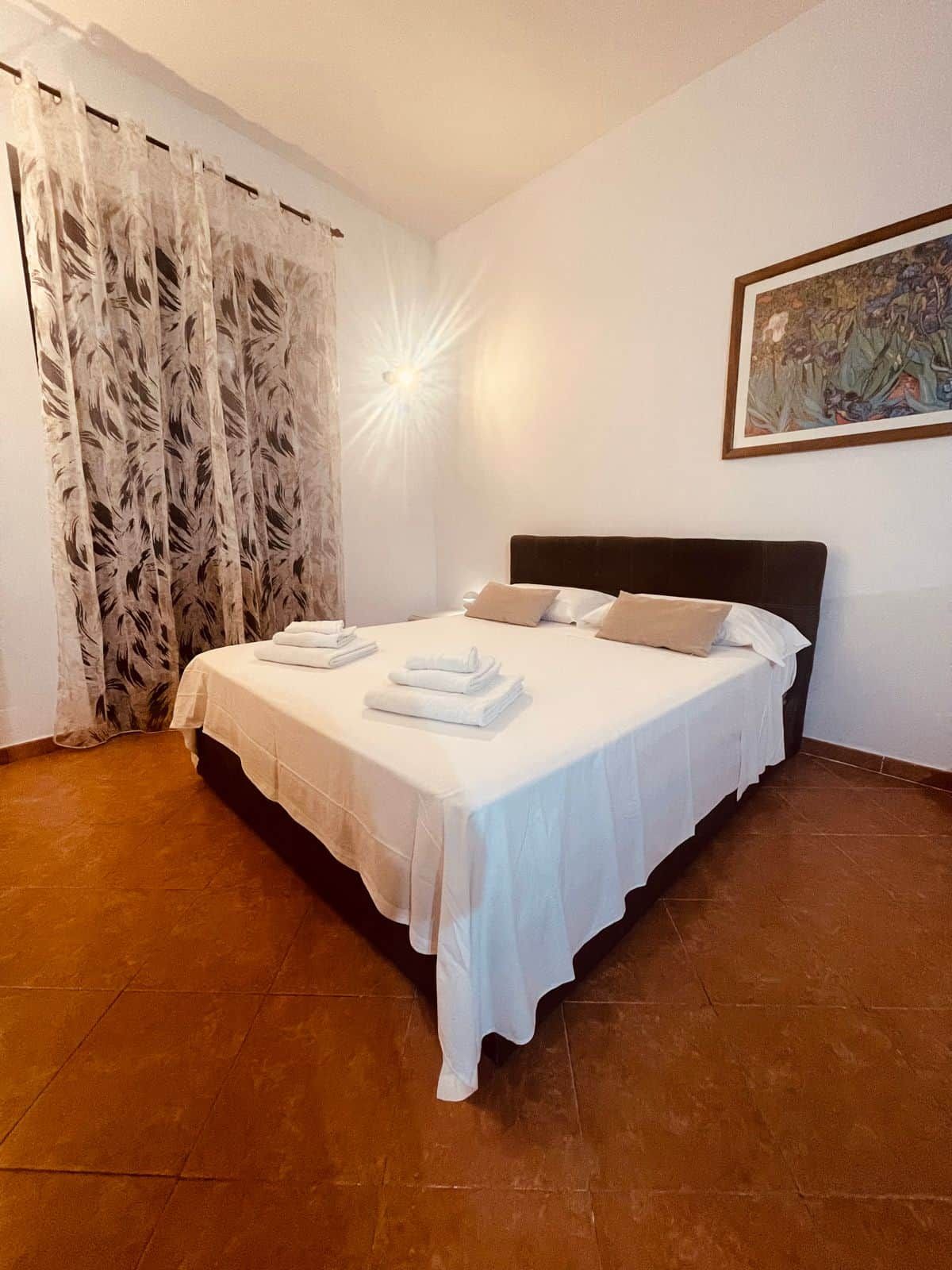 Une chambre d’hôtel avec un lit, des rideaux blancs, un tableau au mur et un sol carrelé.