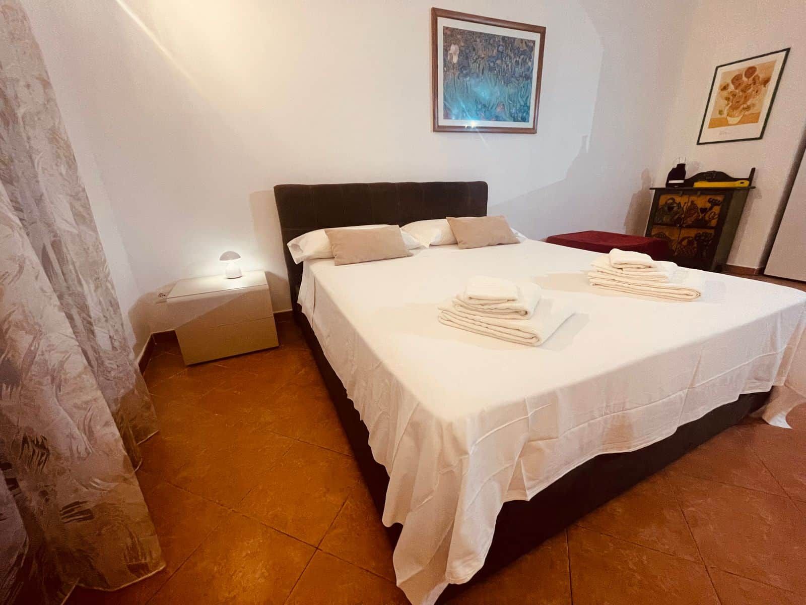 A neatly made bed with pillows and folded towels, a bedside table with a lamp, and artwork on the walls.