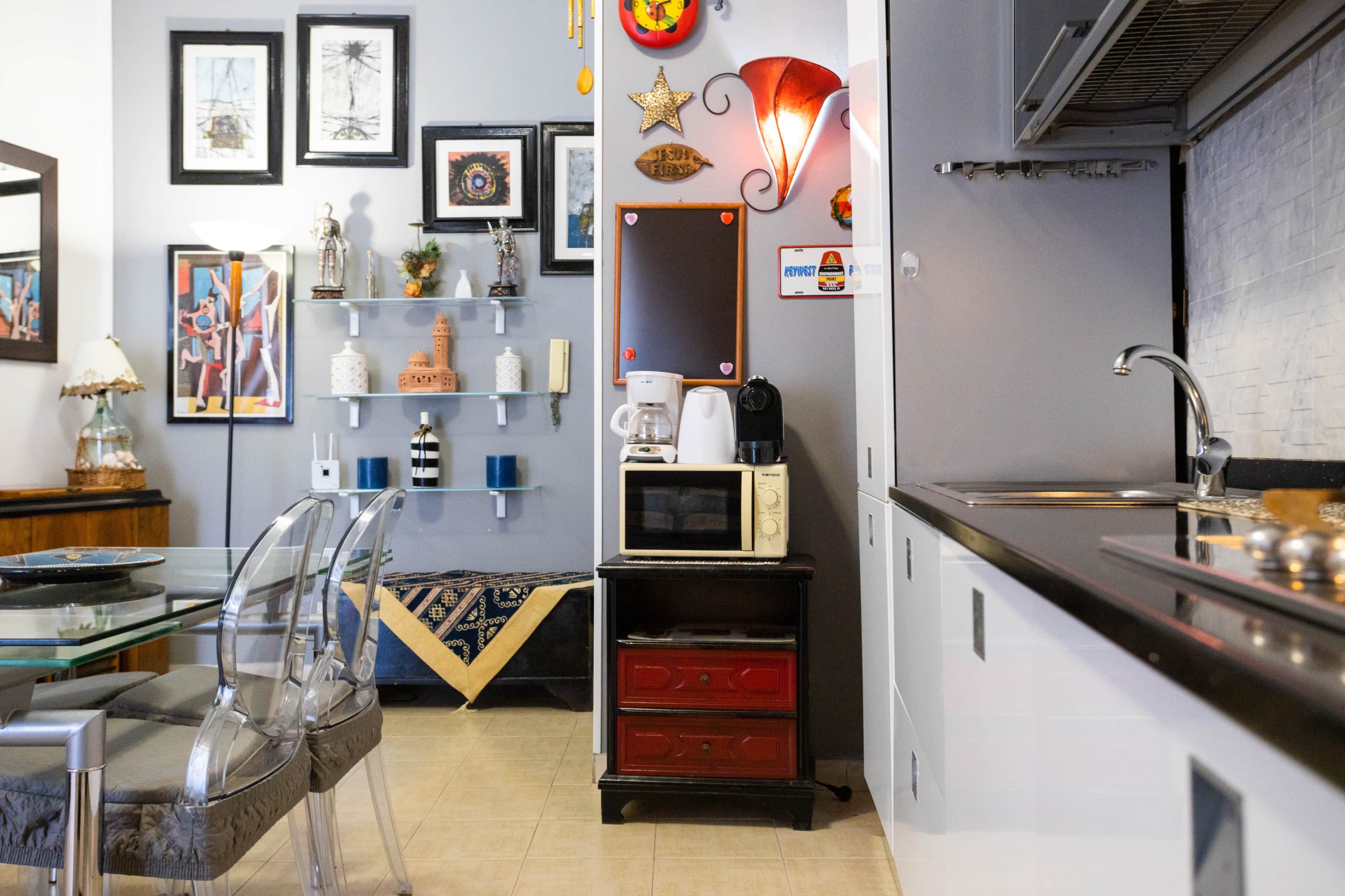 Une cuisine équipée d’un four à micro-ondes, d’une machine à café et d’un meuble rouge. Les murs sont ornés de tableaux encadrés, et on y trouve une table en verre avec des chaises.