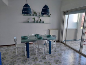 A dining area with a glass table, white chairs, blue accents, and a view through a glass door. - Hola House Mondello Addaura (Palermo)