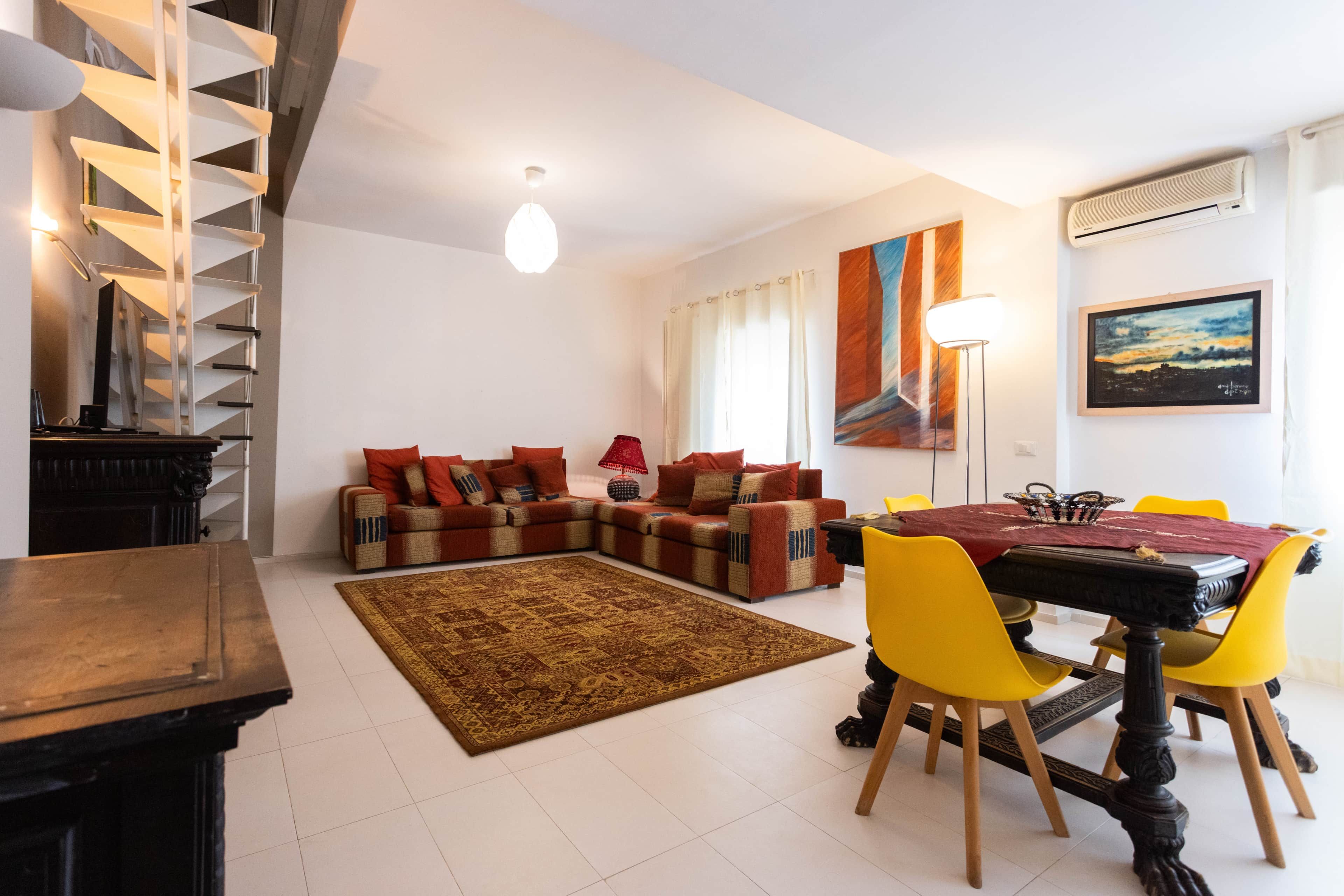 Un salon moderne comprenant un ensemble de canapés, une table à manger avec des chaises, un tapis à motifs et un escalier.