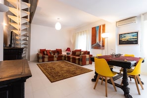 Ein modernes Wohnzimmer mit einer Sofagarnitur, einem Esstisch mit Stühlen, einem gemusterten Teppich und einer Treppe.