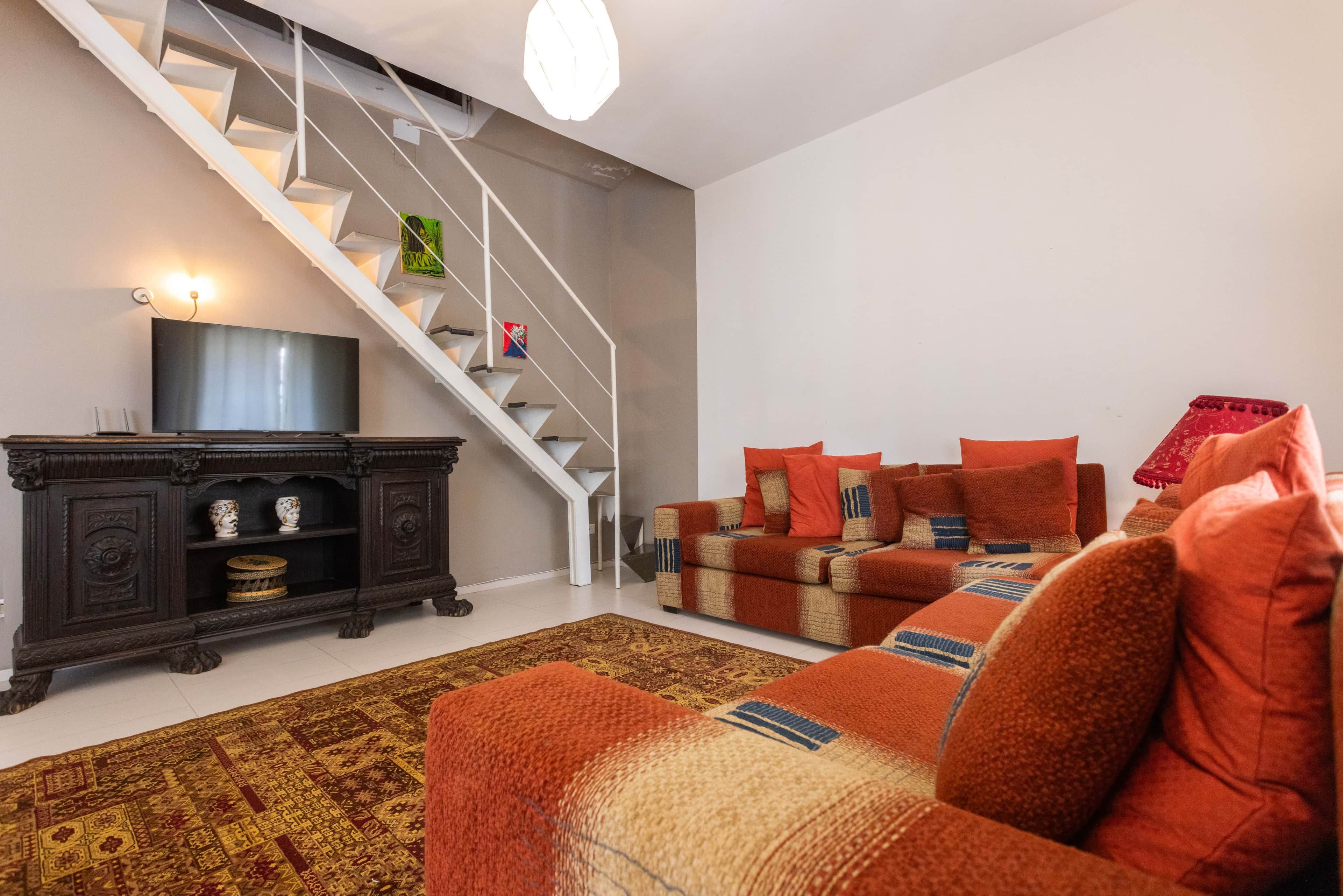 Un salon avec un escalier, un téléviseur, un meuble en bois foncé, un tapis à motifs et un canapé agrémenté de plusieurs coussins.