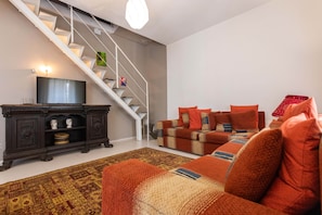 Ein Wohnzimmer mit Treppe, Fernseher, einem dunklen Holzschrank, einem gemusterten Teppich und einer Couch mit vielen Kissen.