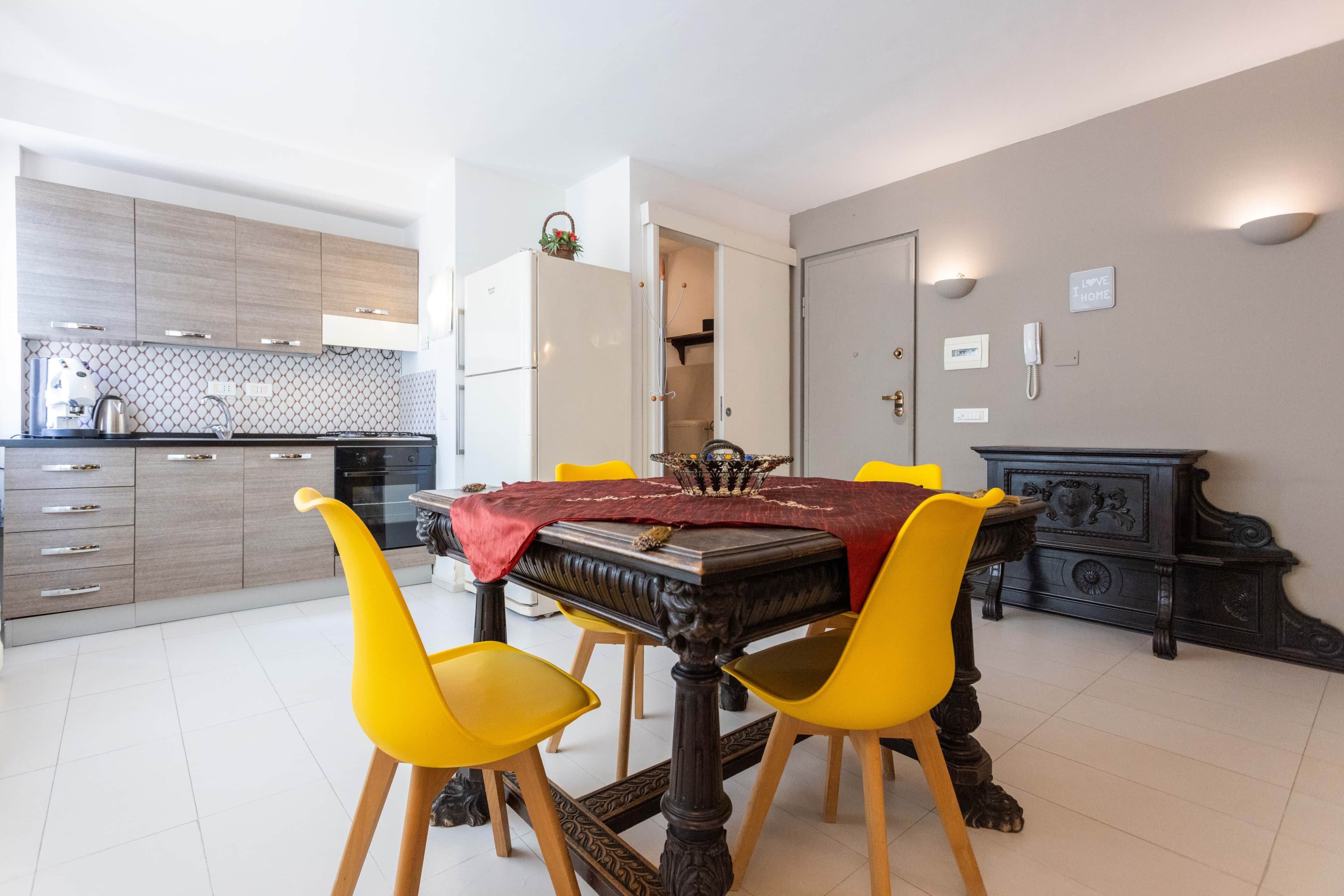 Un coin repas avec une table dressée pour quatre personnes, entourée de chaises jaunes. Une nappe rouge recouvre la table, et un élément décoratif est placé au centre. Au fond de la pièce, on aperçoit une cuisine moderne avec des meubles en bois et un pare-feu en carrelage.