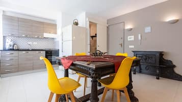 Un coin repas avec une table dressée pour quatre personnes, entourée de chaises jaunes. Une nappe rouge recouvre la table, et un élément décoratif est placé au centre. Au fond de la pièce, on aperçoit une cuisine moderne avec des meubles en bois et un pare-feu en carrelage.