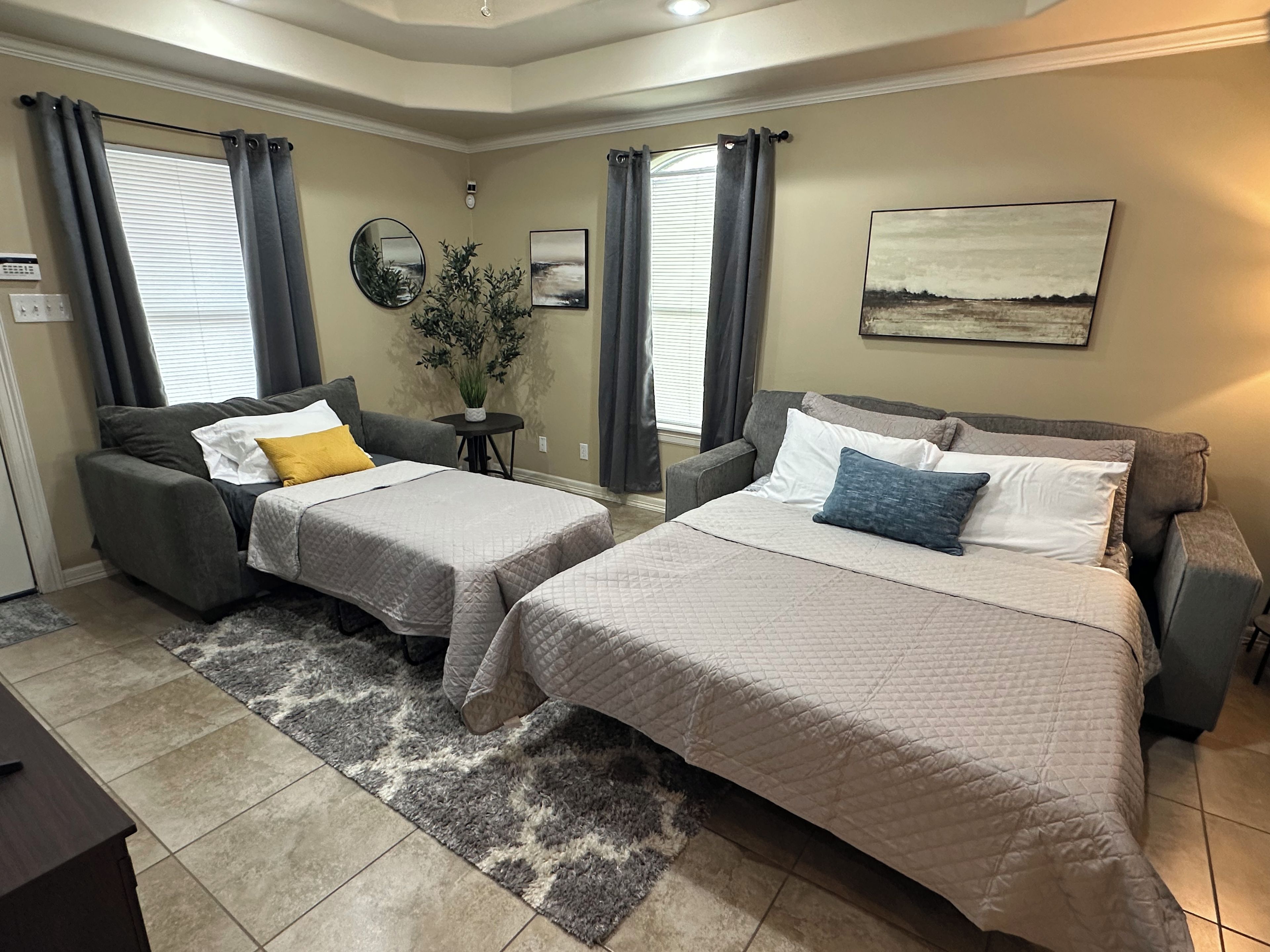 A hotel room with two beds, a gray sofa, a small table with a plant, and a painting on the wall.