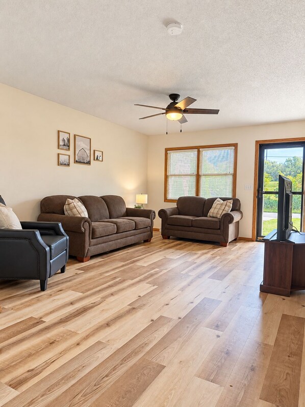 A spacious living room with hardwood flooring, two brown sofas, a black armchair, a ceiling fan, and a large window with blinds.