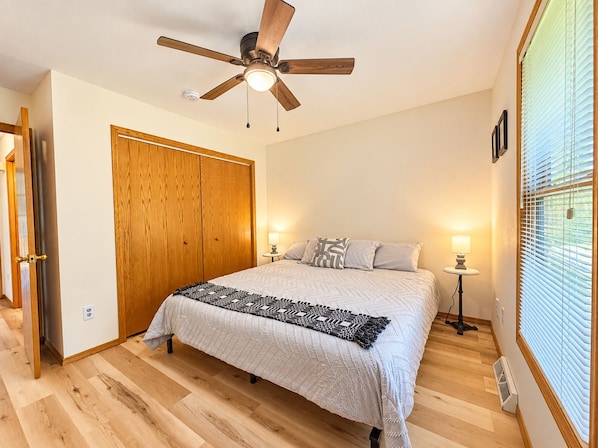 Un dormitorio con cama, ventilador de techo, armario de madera, lámparas de noche y ventana con persianas.