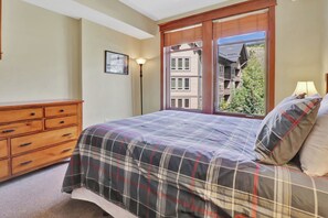 A bedroom with a bed, a wooden dresser, a standing lamp, and a window with a view of buildings and trees. - Lake and Mountain Views. Center Village Gem. In unit washer and dryer! Steps fro (Copper Mountain)