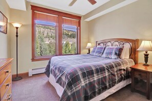 A bedroom with a bed, two bedside lamps, a nightstand, a dresser, and a window with a view of trees and hills. - Lake and Mountain Views. Center Village Gem. In unit washer and dryer! Steps fro (Copper Mountain)