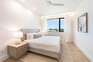 A modern bedroom with a bed, bedside tables, a ceiling fan, a window with curtains, and a city view. - The Sands #5 (Seven Mile Beach)